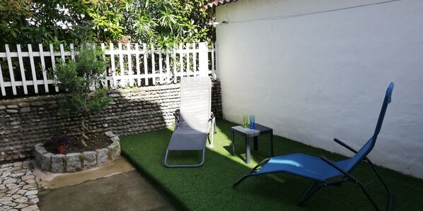 A small outdoor area with artificial grass, a white fence, a stone wall, and a blue chair.
