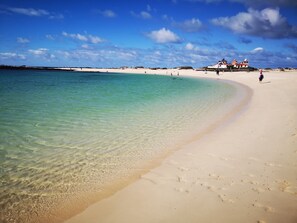 Uma praia com águas turquesa cristalinas, uma faixa de areia e algumas pessoas caminhando.