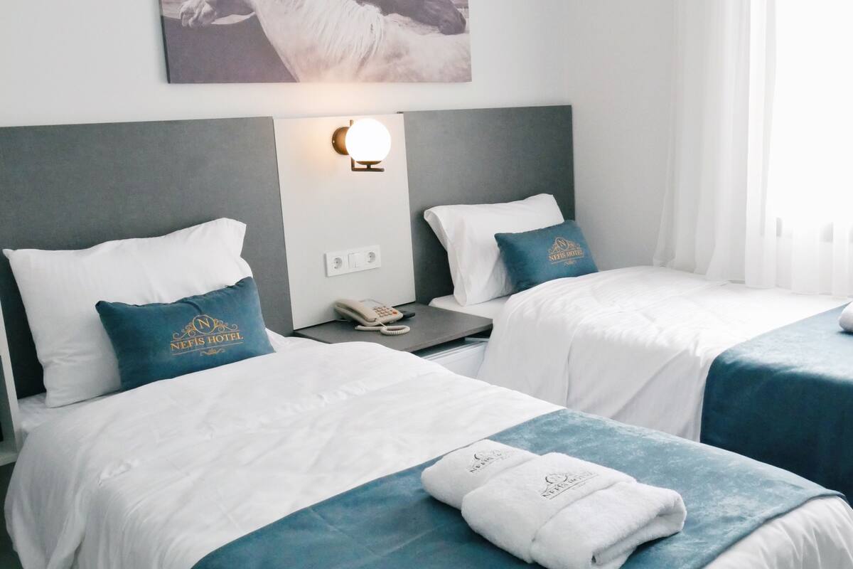 A hotel room with two beds, each with a pillow and a folded towel. There is a wall-mounted light fixture above the beds and a phone on the nightstand.