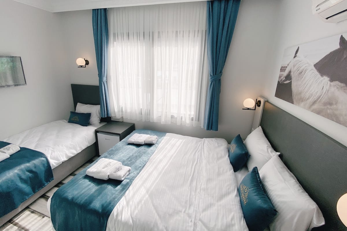 A hotel room with two beds, a nightstand, a wall-mounted lamp, and a large window with blue curtains.