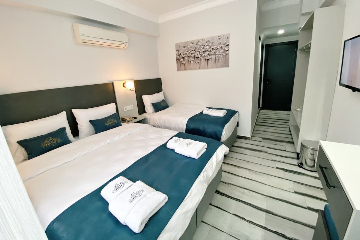 A hotel room with two beds, each with white linens and teal bedspreads. There is a wall-mounted air conditioner and a painting above the beds.