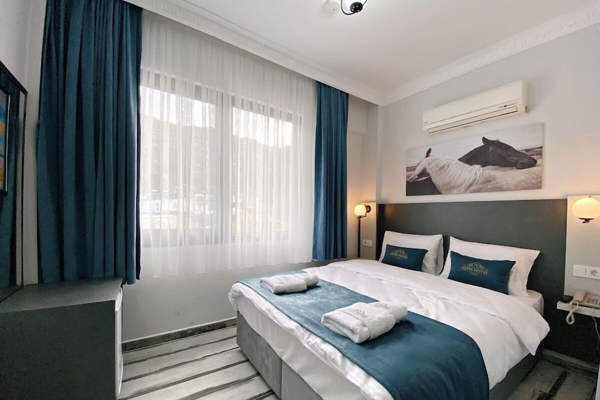 A hotel room with a bed, a desk, a chair, a window with curtains, a wall-mounted air conditioner, and a painting.