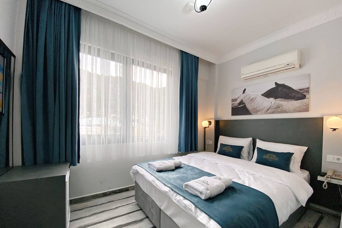 A hotel room with a bed, a desk, a chair, a window with curtains, an air conditioning unit, a wall-mounted phone, and a picture of a horse.