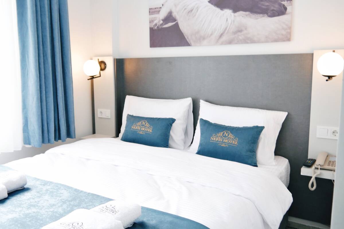 A hotel room with a large bed, two pillows with the hotel's logo, a bedside phone, and a window with curtains.