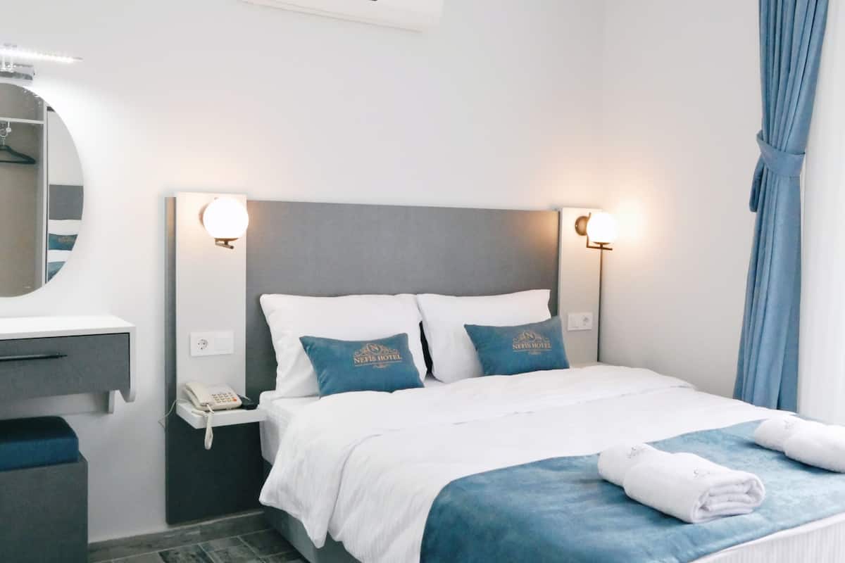 A hotel room with a bed, a bedside table with a phone, a mirror, and a wall-mounted light fixture.