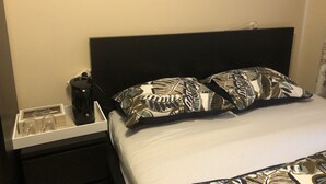 Ein ordentlich bezogenes Bett mit gemusterten Kissen, ein schwarzes Kopfteil und ein Nachttisch mit Lampe und Taschentuchbox.