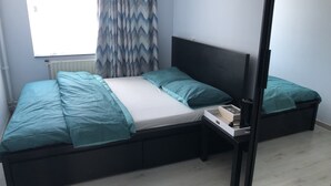 Ein Einzelbett mit schwarzem Gestell und weißer Matratze, ein Nachttisch mit einem Buch und Kopfhörern sowie ein Fenster mit Vorhang.
