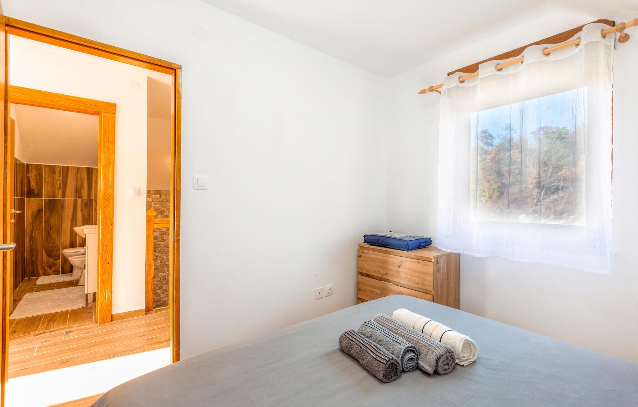 A bedroom with a bed, a window with a view, a wooden dresser, and a bathroom visible through an open door.