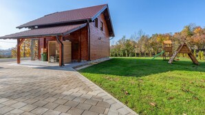 A wooden cabin with a tiled roof, a covered porch, and a grassy yard with a wooden play structure. - Amazing home in Perusic with sauna (Perusic)