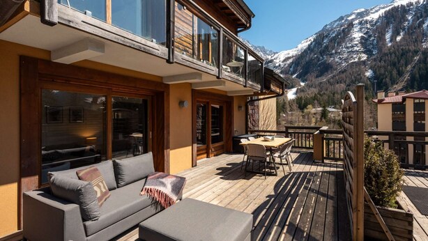 Un terrazzo in legno con mobili da esterno, una porta vetrata che dà su una stanza e vista sulle montagne.