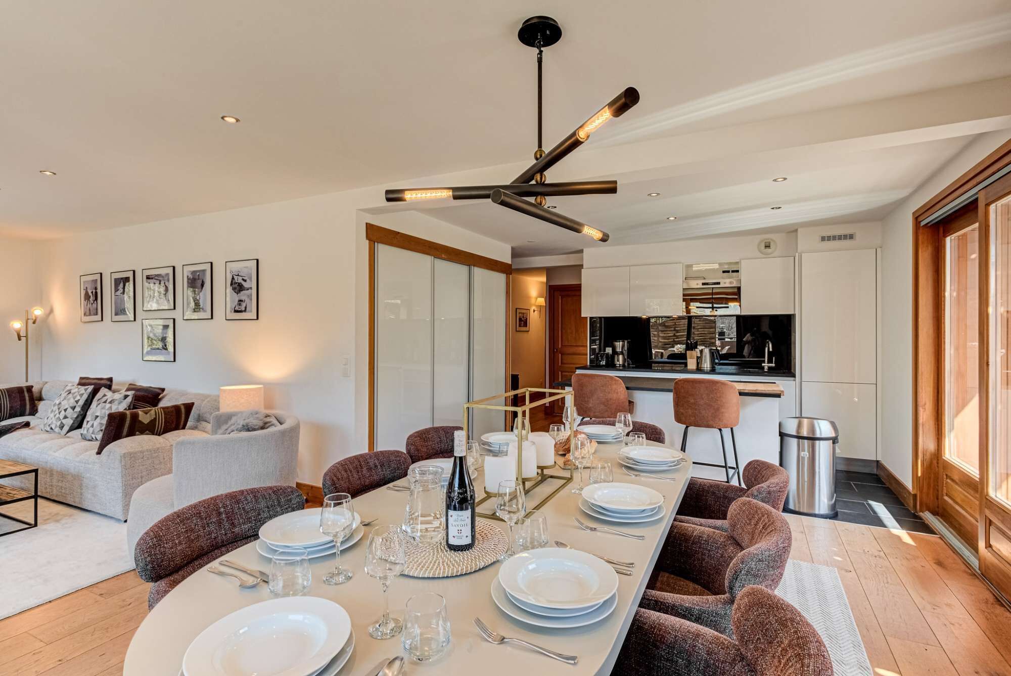 Un comedor moderno con una larga mesa dispuesta para seis personas, una cocina al fondo y una sala de estar con un sofá y cojines.