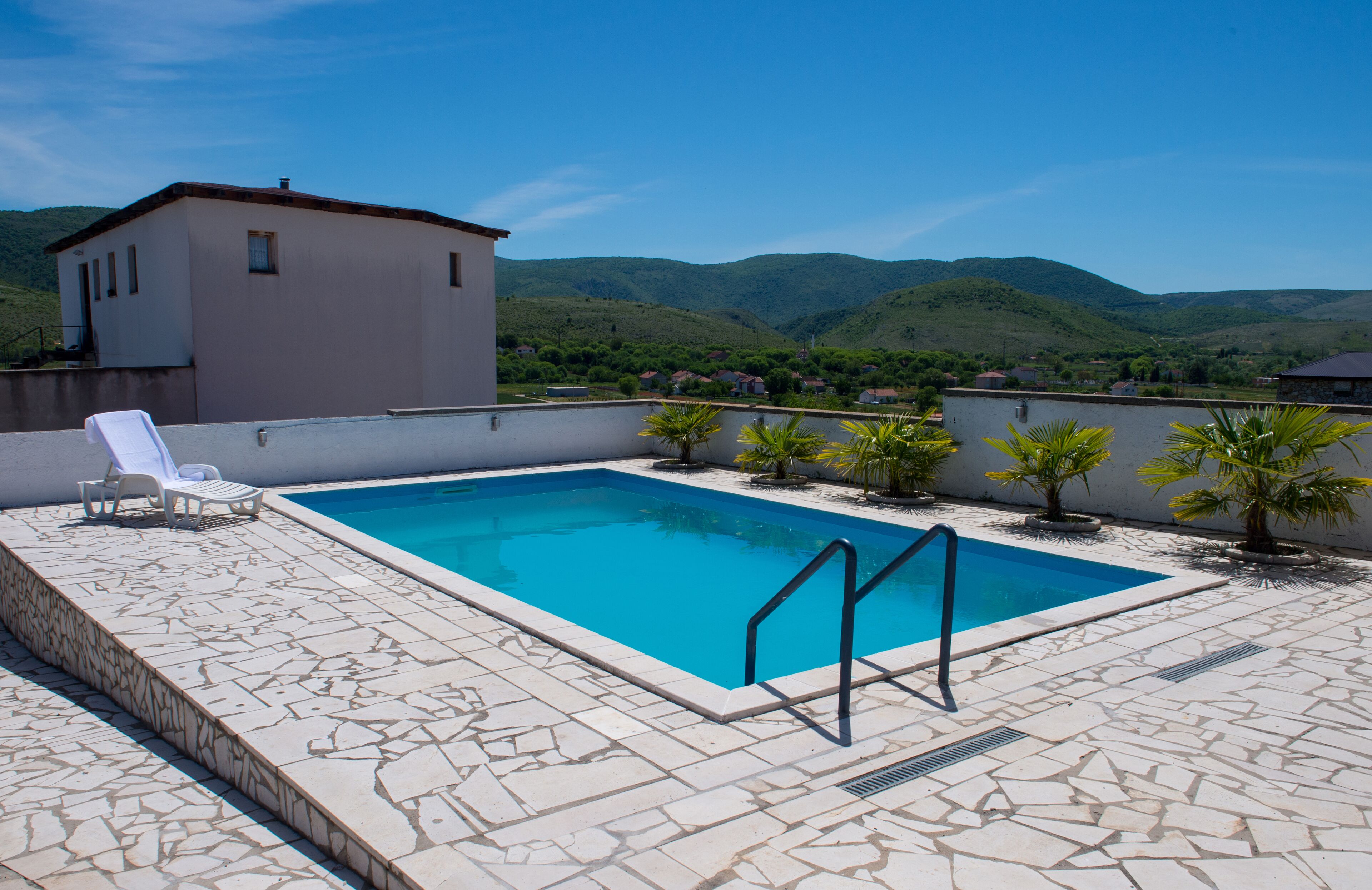Una piscina rectangular con un deck con baldosas de piedra, un edificio blanco al fondo y un paisaje montañoso.