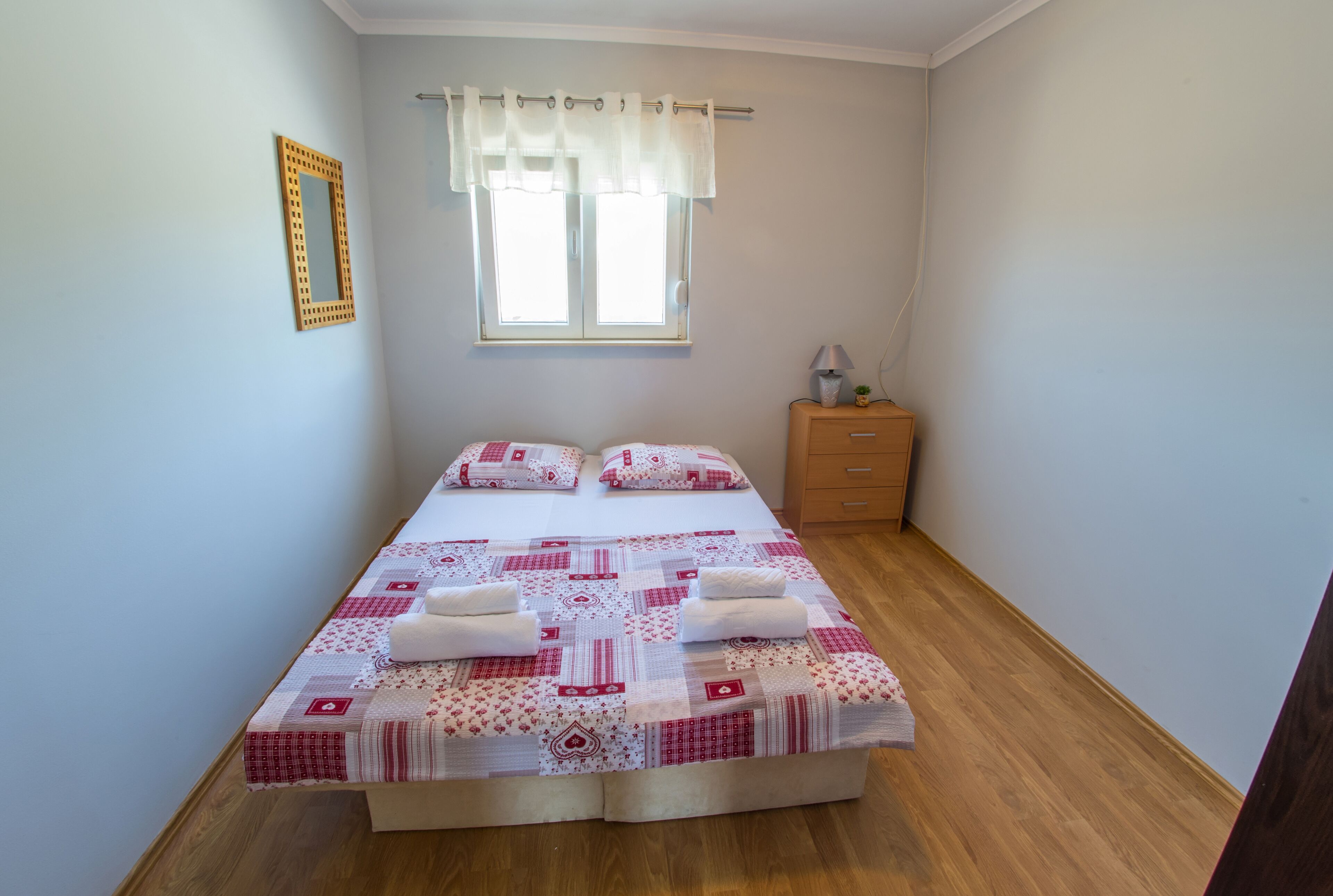 A small, neatly arranged bedroom with a bed, pillows, a nightstand, and a window with curtains.