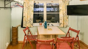 A room with a wooden table and four red chairs, a window with floral curtains, a television on the wall, and a barrel in the corner.