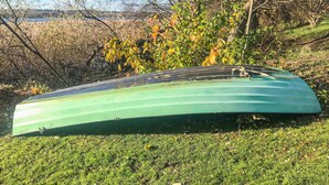A green boat lying on grass with a background of trees and a body of water.