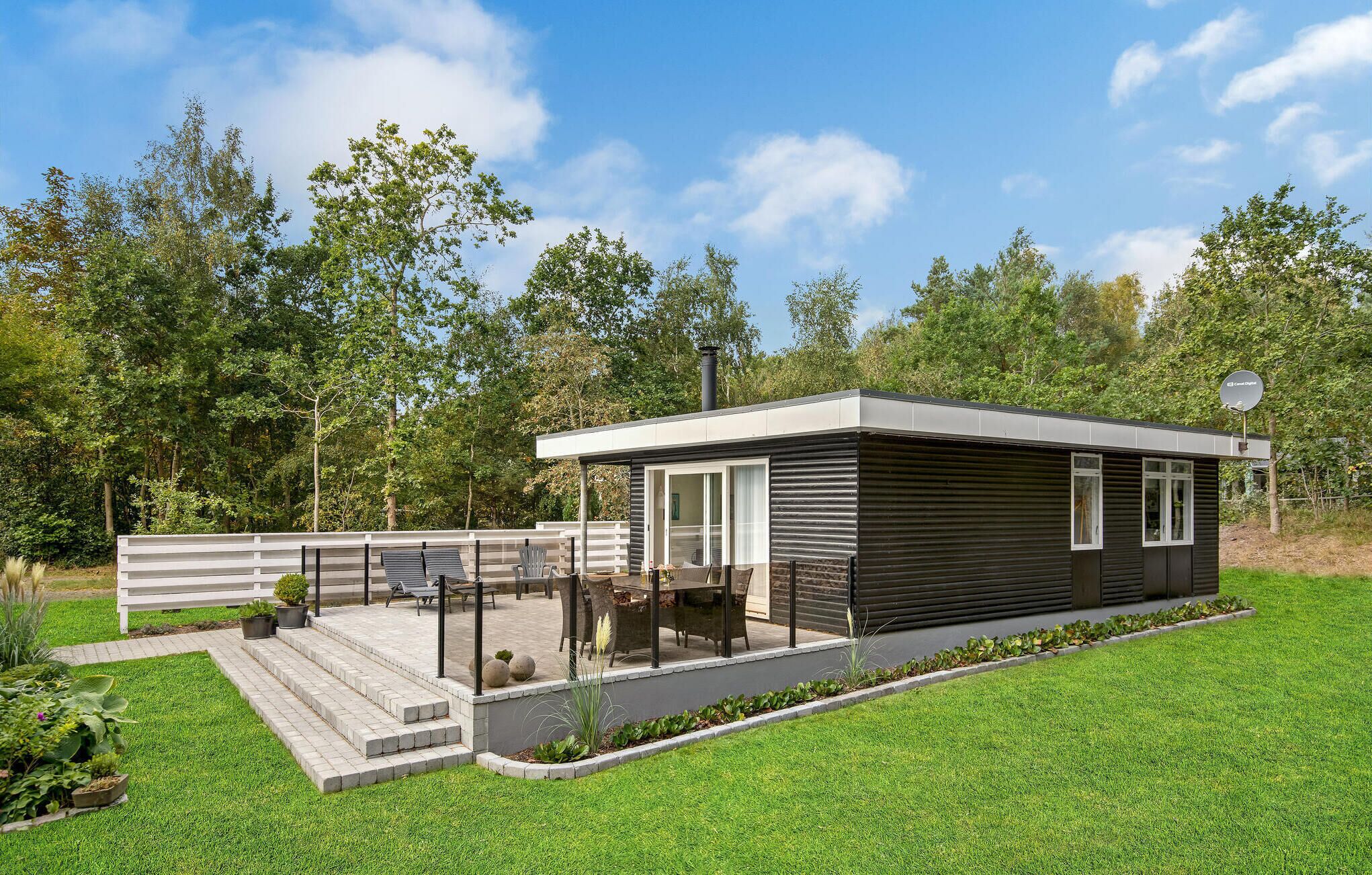 A modern, single-story cabin with a covered patio, surrounded by lush greenery and a clear sky.