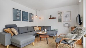 A modern living room with a grey sectional sofa, a wicker chair, and a round coffee table. There are wall art pieces and a television on the wall. - Beautiful home in Aakirkeby with WiFi (Aakirkeby)