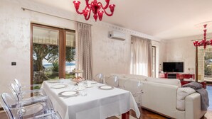 Zona pranzo con tavolo apparecchiato per quattro, sedie trasparenti, un divano bianco e una televisione sullo sfondo.
