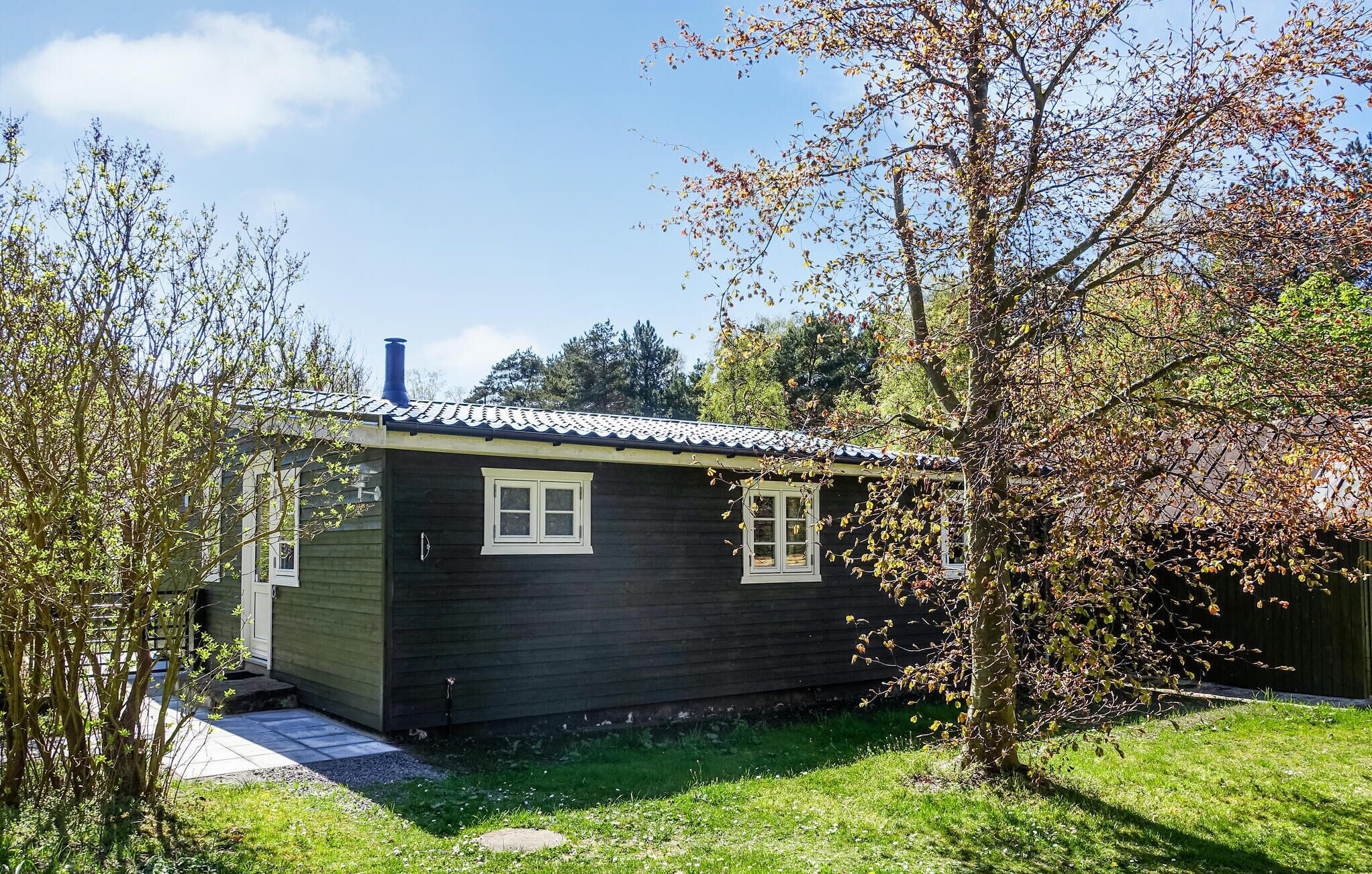 Une petite cabane en bois sombre, avec un toit recouvert de tuiles, entourée de verdure et d’arbres.