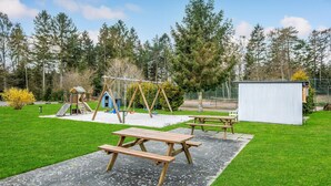 Une aire de jeux avec des balançoires et des toboggans, une table de pique-nique et un abri dans un espace verdoyant, avec des arbres en arrière-plan.