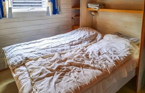 Un lit simple avec une couette blanche, dans une pièce aux murs en bois et dotée d’une fenêtre avec des stores.
