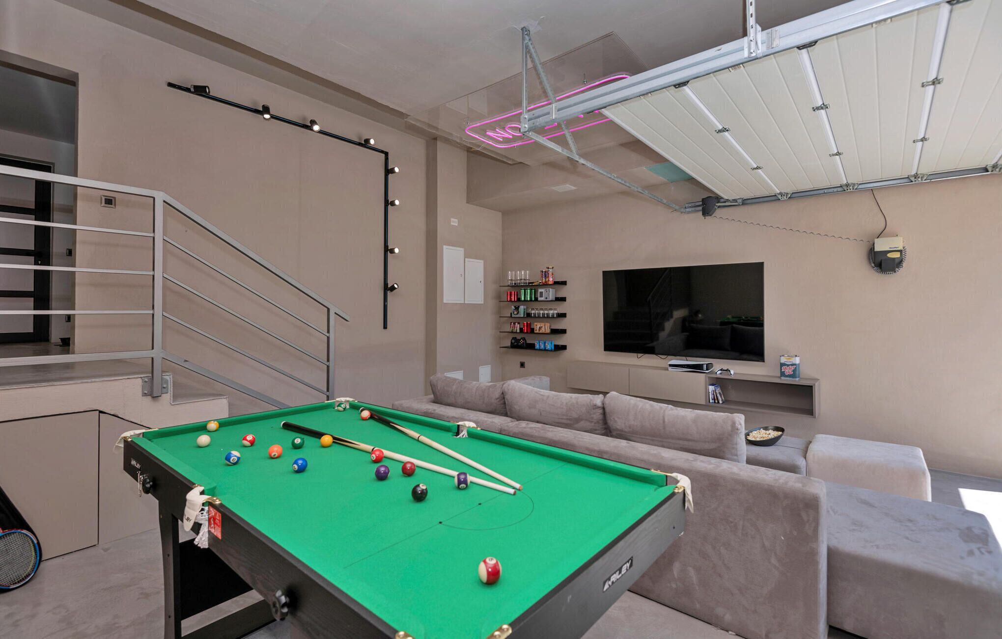 A modern living room with a pool table, a staircase, a flat-screen TV, and a variety of shelves with items.