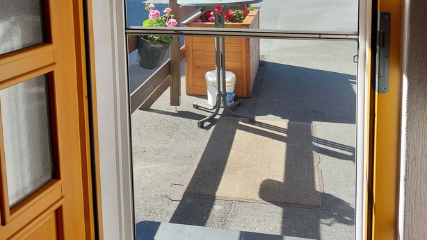 A view through a door with a wooden frame and glass panels, leading to an outdoor area with a covered seating area, a wooden bench, and a planter with flowers.