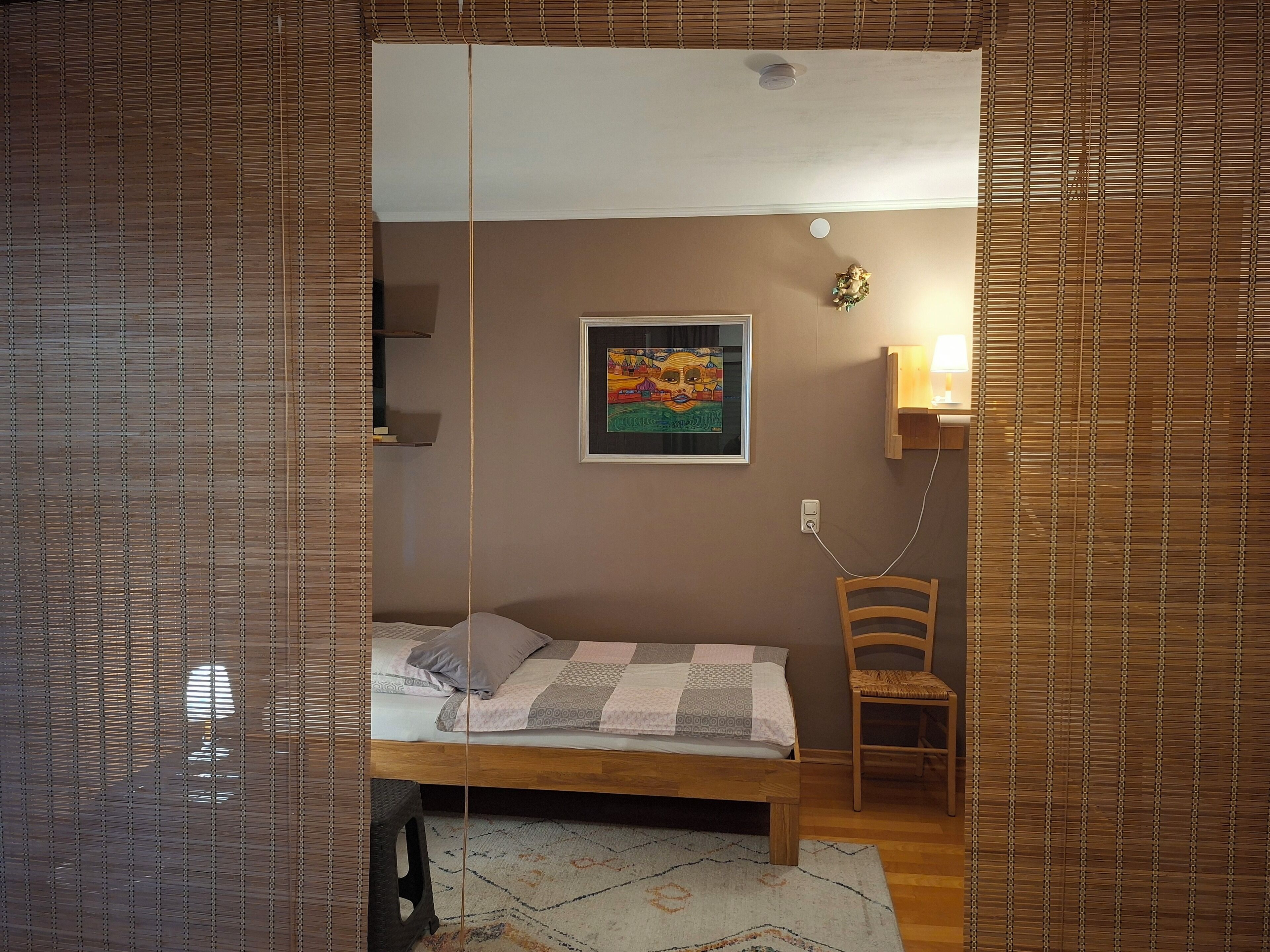 A bedroom with a bed, a chair, a small table with a lamp, and a framed picture on the wall.