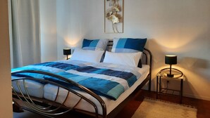 A neatly made bed with a blue and white quilt, two bedside lamps, a framed artwork on the wall, and a white curtain.