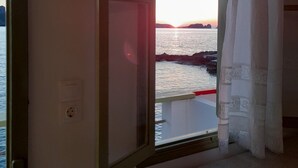 A room with a bed, a window with an open curtain, and a view of a sunset over water. - Milos By The Sea – Seafront House (Milos)