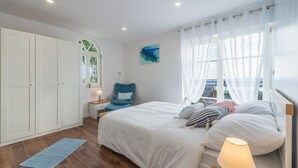 Un dormitorio con cama, una silla, un armario, una ventana con cortinas y un cuadro en la pared.