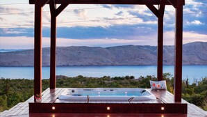 Terraza de madera con un jacuzzi y una copa de vino tinto, con vistas a un lago y montañas.