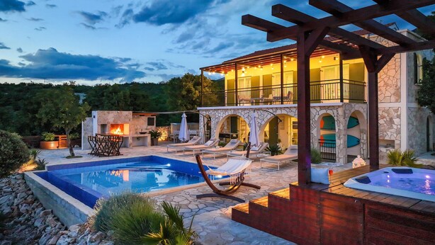 Un área de piscina exterior lujosa con un jacuzzi, sillones de descanso y un edificio de piedra con ventanales de arco.