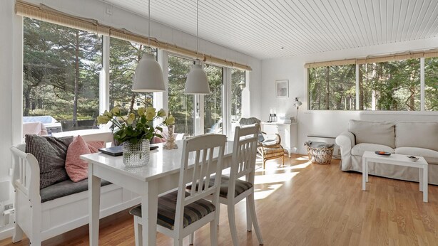 Un intérieur lumineux et blanc, avec de grandes fenêtres, une table à manger, des chaises, un canapé et une petite table d’appoint.