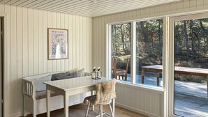Une pièce accueillante avec un banc en bois blanc, une table et une chaise. Un tableau encadré orne le mur, et une fenêtre offre une vue sur les arbres.
