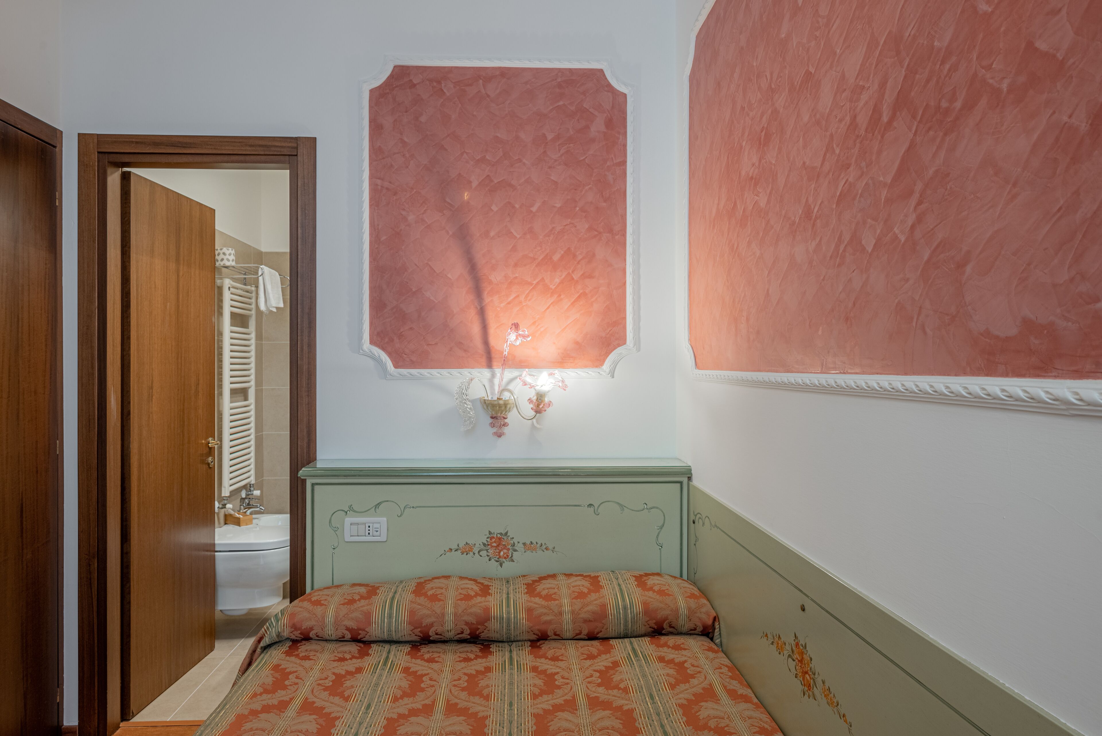 A bedroom with a bed, a nightstand, a wall-mounted light fixture, and a door leading to a bathroom.