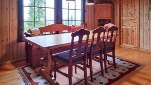 Un comedor de madera con una mesa grande y sillas, una ventana con vista a árboles, y un aparador con cristalería.