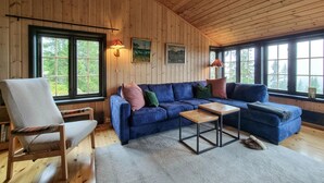 Interior acogedor de una cabaña con un sofá azul, una silla de madera, una mesita y ventanas amplias con vistas a los árboles.