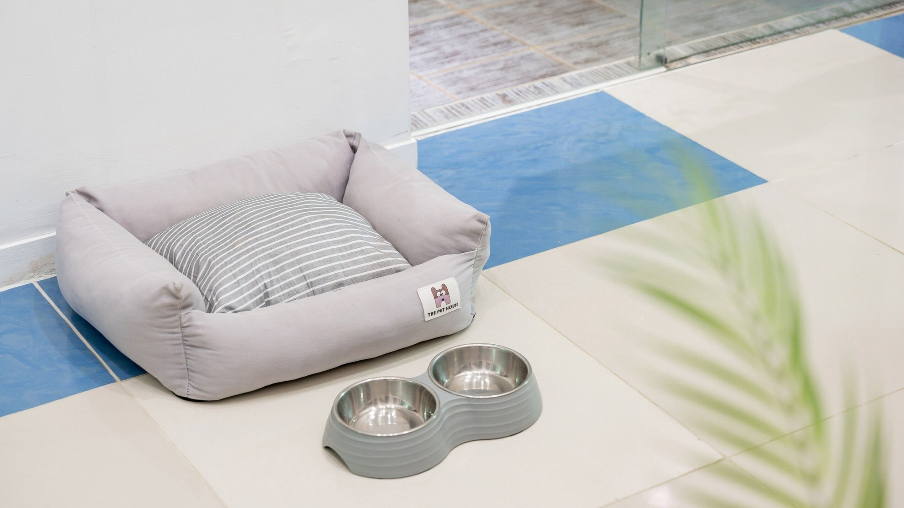 A pet bed with a tag and two metal bowls on a tiled floor.
