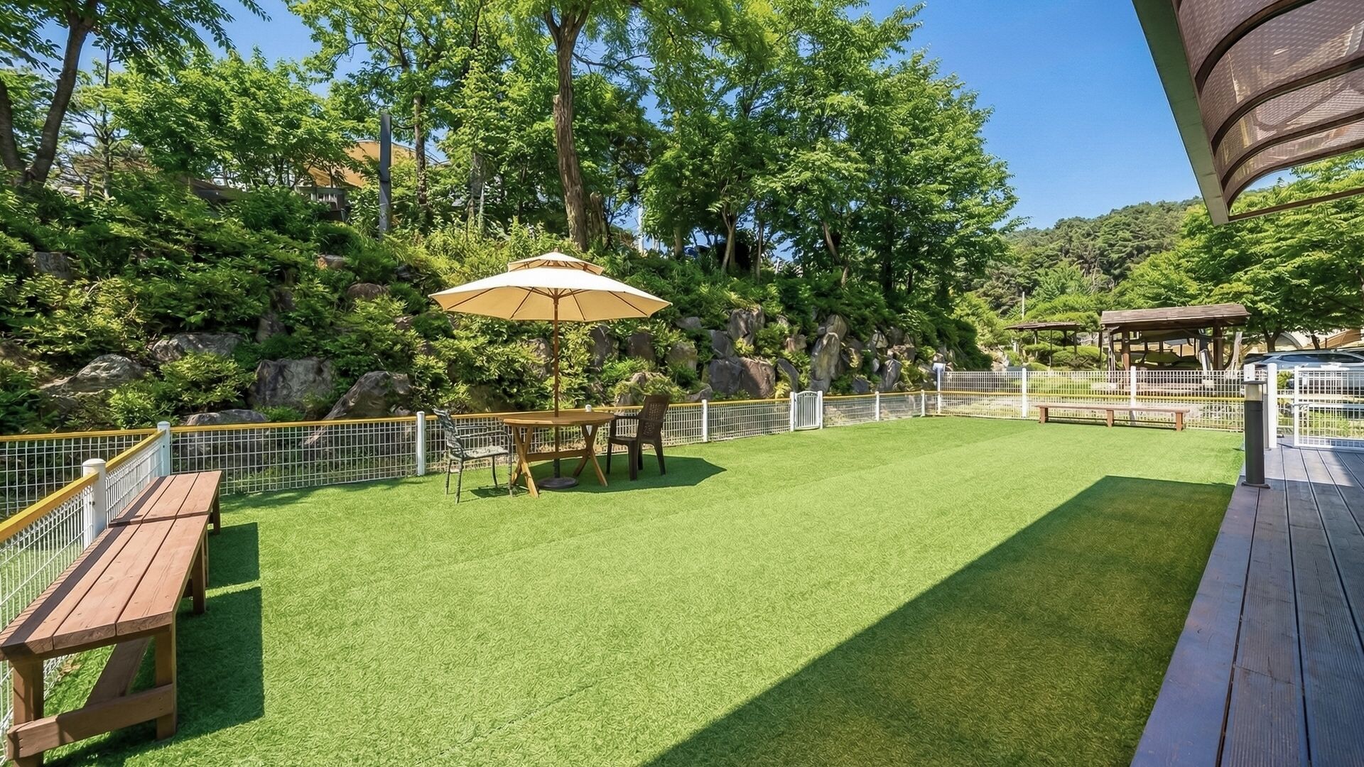 Ein gepflegter Außenbereich mit Kunstgrasanlage, einem Holztisch mit Stühlen, einer Bank und einer Pergola.