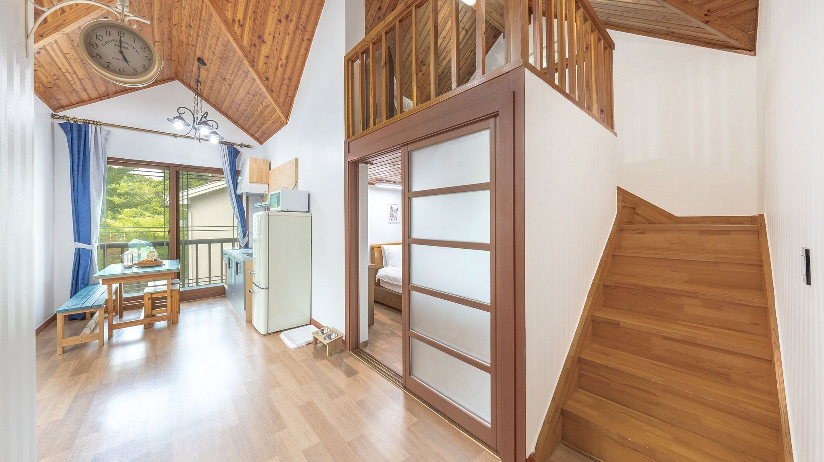A room with a wooden ceiling, a dining area with a table and chairs, a bed with a white headboard, and a sliding door with frosted glass panels.
