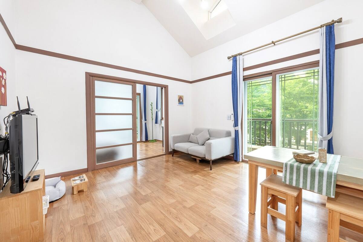 A room with a wooden floor, a sofa, a dining table with chairs, a TV, and a window with curtains.