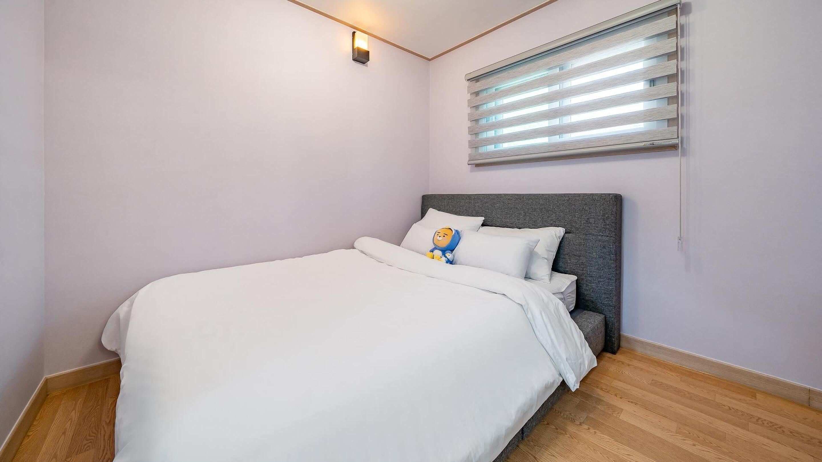 A bedroom with a bed, a nightstand, a wall-mounted light, and a window with blinds.