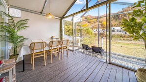 Una terraza de madera con una mesa y sillas, amplias ventanas que ofrecen vistas a un paisaje montañoso y un colgador con luz.