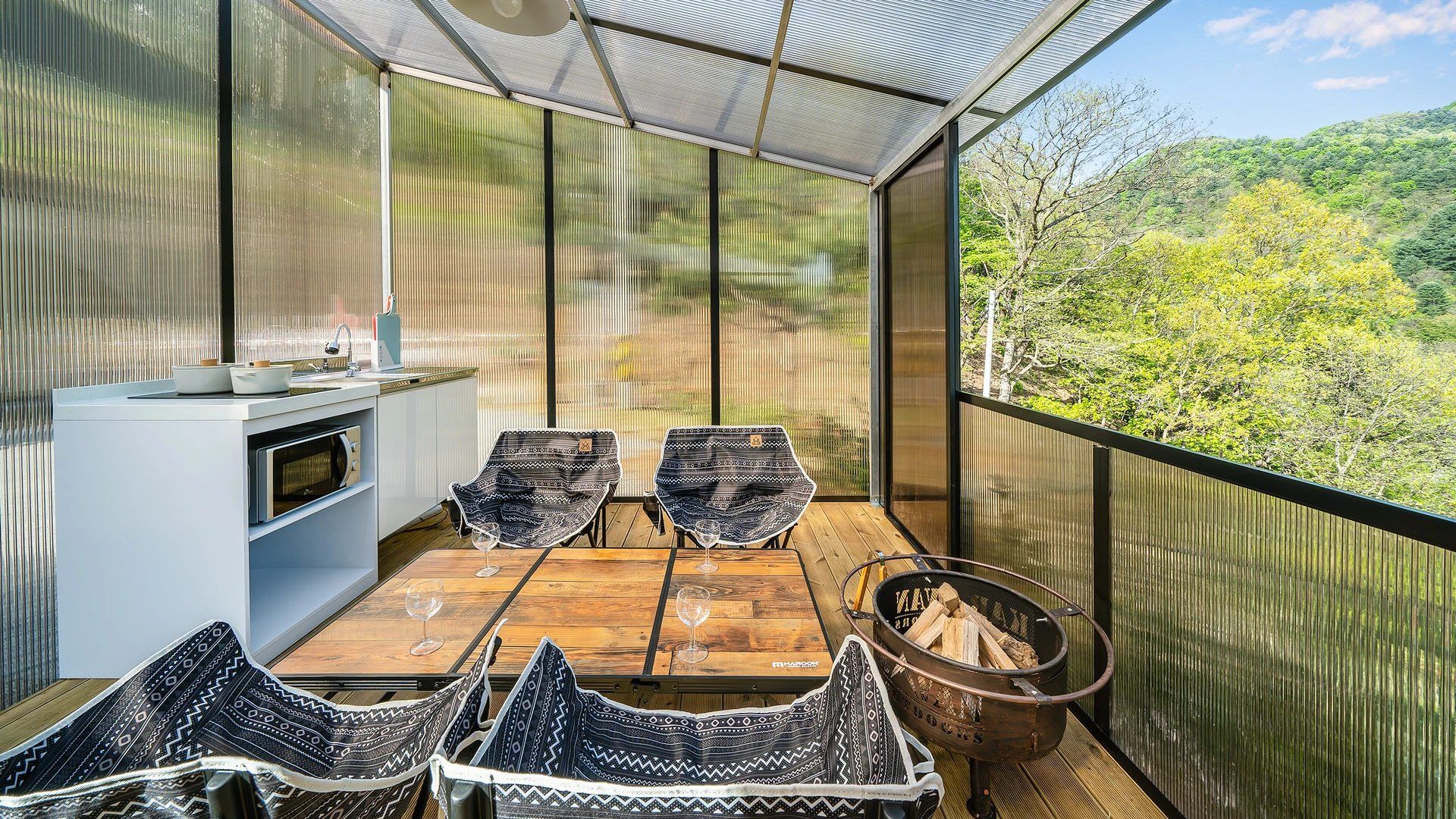 A balcony with a wooden table, two chairs, a small kitchen area, and a view of the surrounding greenery.