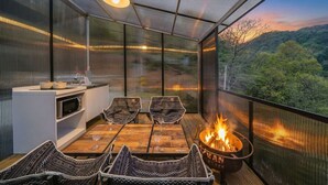 Un balcón acogedor con una fogata, una mesa de comedor y sillas, con vistas a una ladera boscosa.