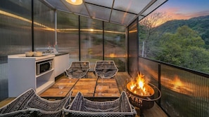 Varanda aconchegante com lareira, mesa de jantar e cadeiras, com vista para uma encosta arborizada.