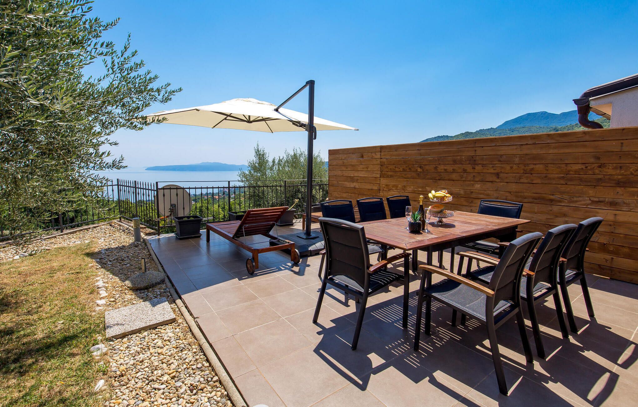 Eine Terrasse im Freien mit Tisch und Stühlen, einem großen Sonnenschirm und Blick aufs Meer.