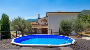 Piscina com deck de madeira, cercada por oliveiras e um edifício com telhado em azulejos.