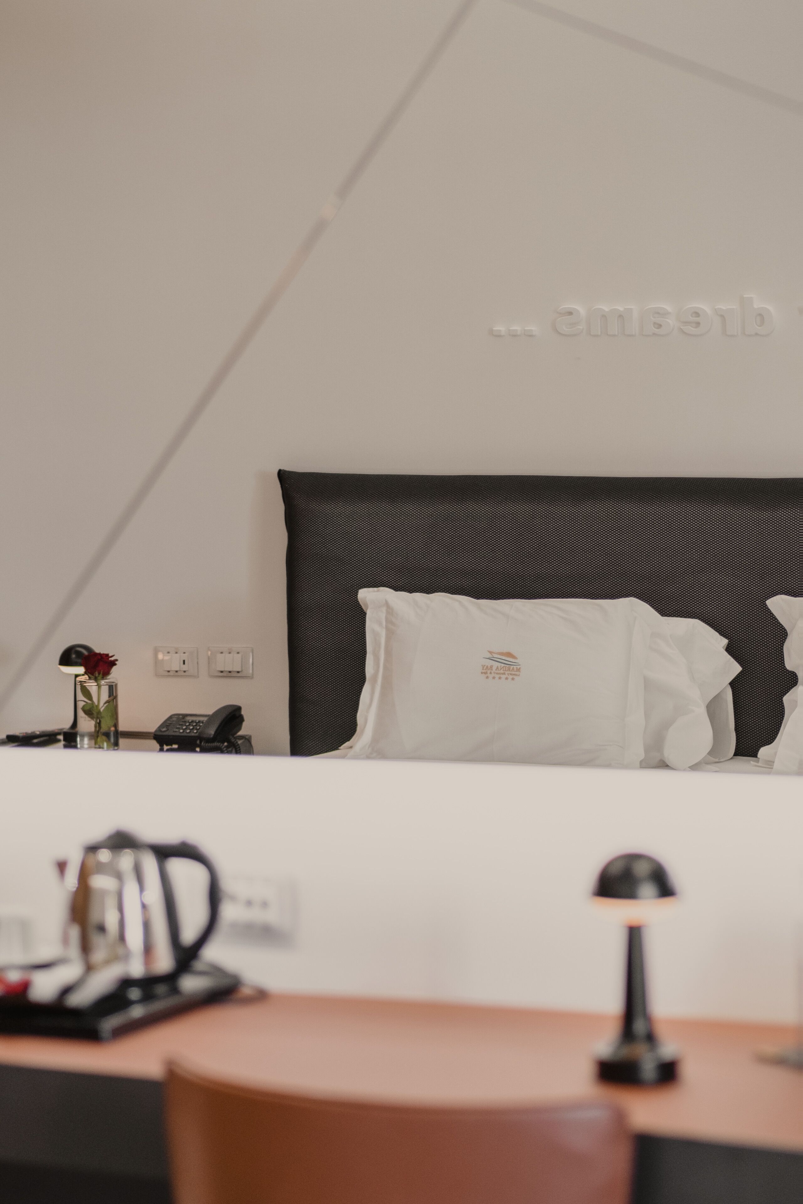 A hotel room with a bed, a bedside table with a lamp, a phone, and a vase with a rose.